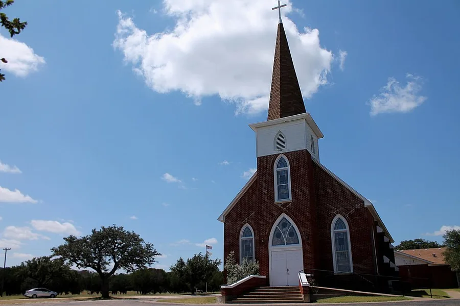 Our Savior's Lutheran Church at Norse, Texas, photographed in 2014: a red-brick chapel with pointed stained-glass windows, a white double door above a short flight of steps, and a tall dark-shingled spire on a white belfry, standing among live oaks under a wide blue Texas sky.