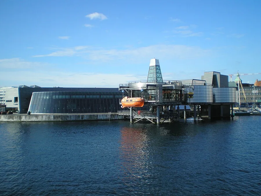 Waterfront exterior of the Norwegian Petroleum Museum in Stavanger: an angular glass-and-steel building beside a tower and platform-like superstructure on pilings over the harbor, with an orange lifeboat suspended alongside, under a clear blue sky.