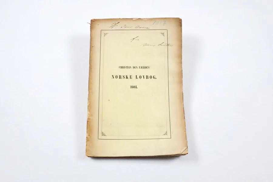 Photograph of a worn, yellowed paper-wrappered copy of the 1604 Norwegian law-book from Ivar Aasen's library, its plain cover printed with the title 'Christian den Fjerdes Norske Lovbog. 1604.' inside a decorative border, with old handwritten ownership notes above.