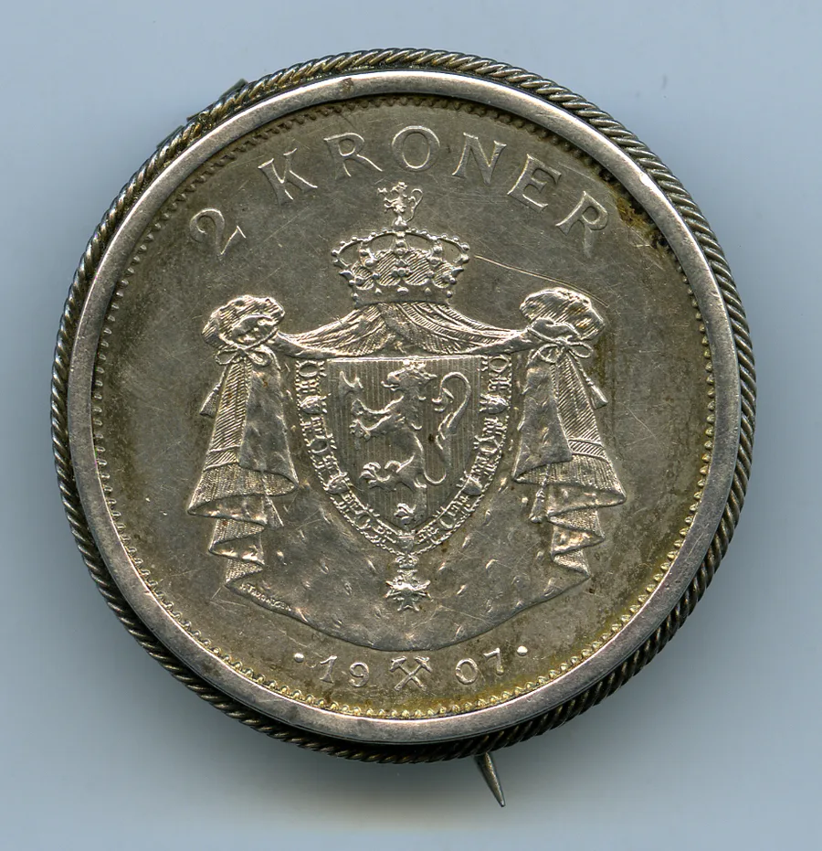 Obverse of a worn silver Norwegian 2-kroner coin: a crowned shield bearing the lion within a draped ermine mantle gathered in two tassel-tied bundles, '2 KRONER' arched above and the date 19·07 with a crossed-hammers mint mark below; set in a twisted-rope brooch mount.