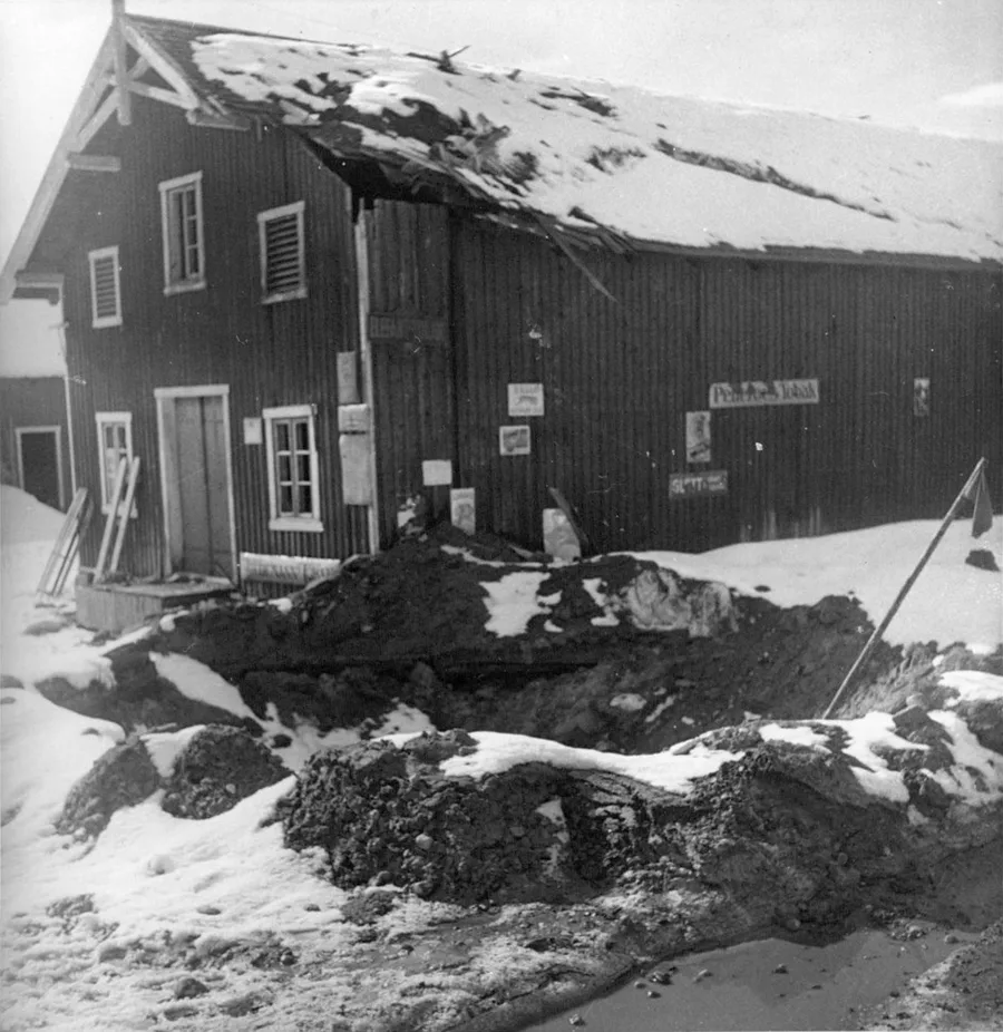 Black-and-white 1940 photograph of a large bomb crater gouged into snowy ground beside a wooden building in Nybergsund, its roof torn open by the blast.