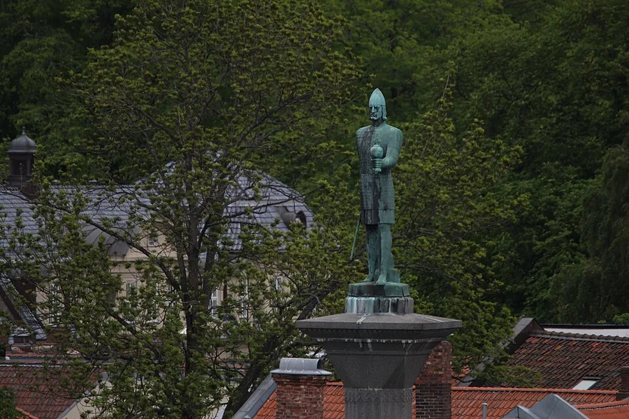 A weathered green bronze statue of a helmeted Viking king in mail armor, sword at his hip, standing atop a tall stone column above red-tiled rooftops and trees in Trondheim.