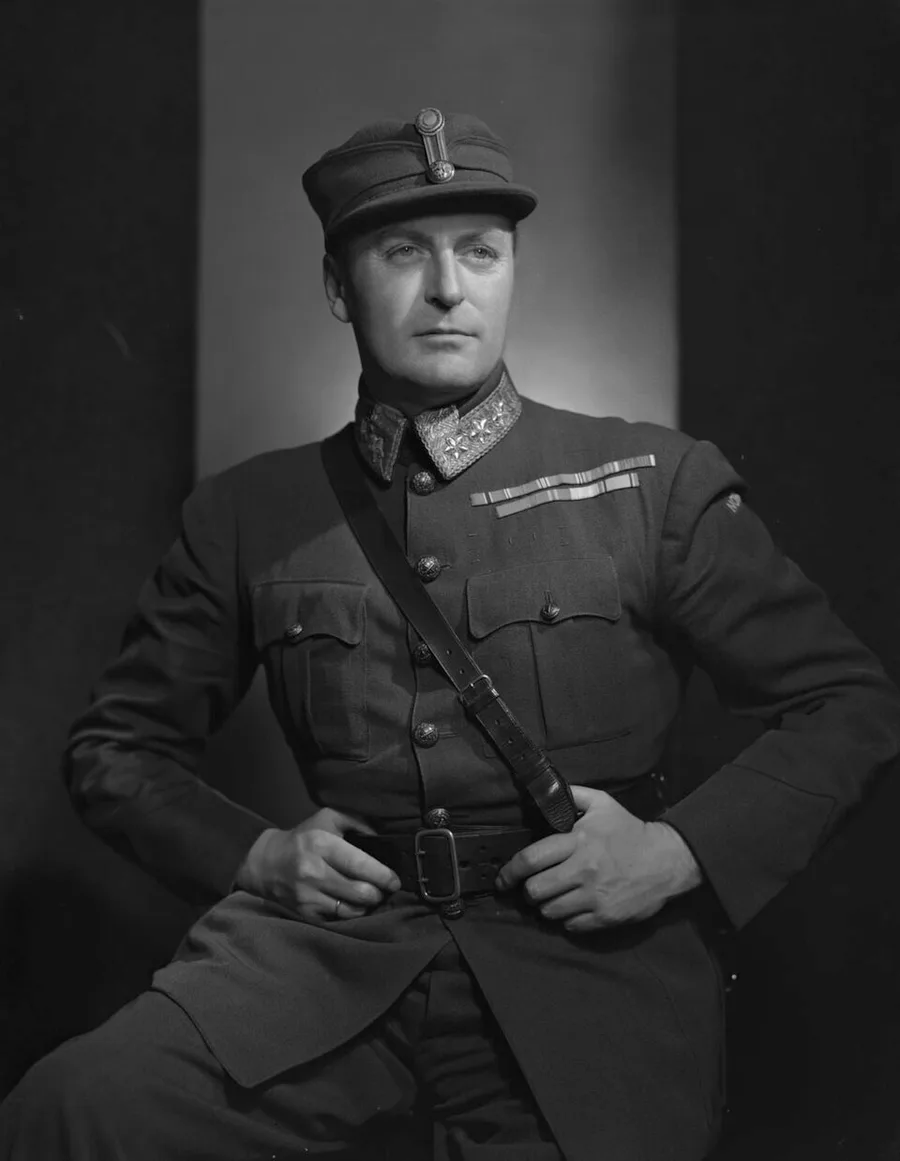 Black-and-white Karsh studio portrait of a young Crown Prince Olav of Norway in military uniform and peaked cap, seated with hands on his belt, photographed against a plain dark backdrop.