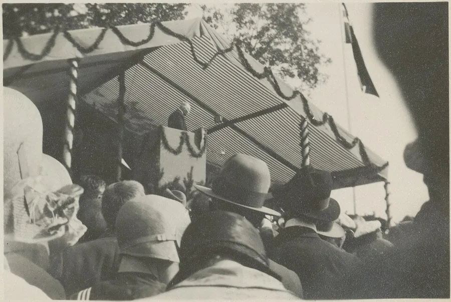 Black-and-white 1930 photograph of a hat-wearing crowd at Stiklestad on Olsok day, listening to a lone speaker at a lectern beneath a garland-swagged striped awning, a flag flying from a pole at the right.