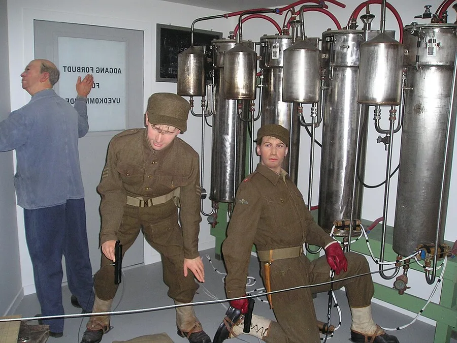 Museum reconstruction display: two mannequins in period military uniform crouched with weapons among tall steel electrolysis cells and red piping, staging the Vemork sabotage; a third figure — the plant's night watchman, hands raised — stands in the background.