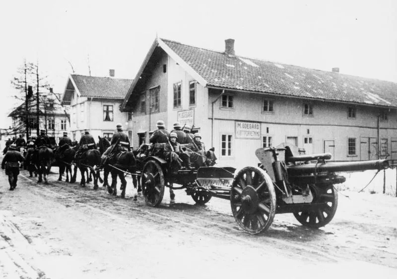 Black-and-white 1940 photograph of German soldiers in helmets leading a horse-drawn artillery piece down a snow-covered street past houses near Oslo.