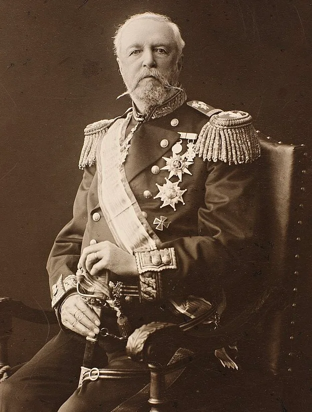 Sepia photographic portrait of an elderly white-bearded man seated in a chair, wearing a military uniform with epaulettes, a sash, medals, and a sword — King Oscar II of Sweden and Norway, circa 1905.