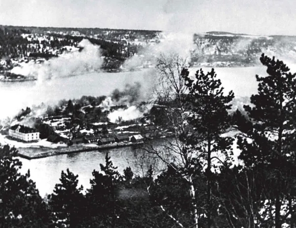 Black-and-white 1940 photograph looking across the Oslofjord through pine boughs at Oscarsborg Fortress, columns of smoke rising along the shoreline during the air attack.