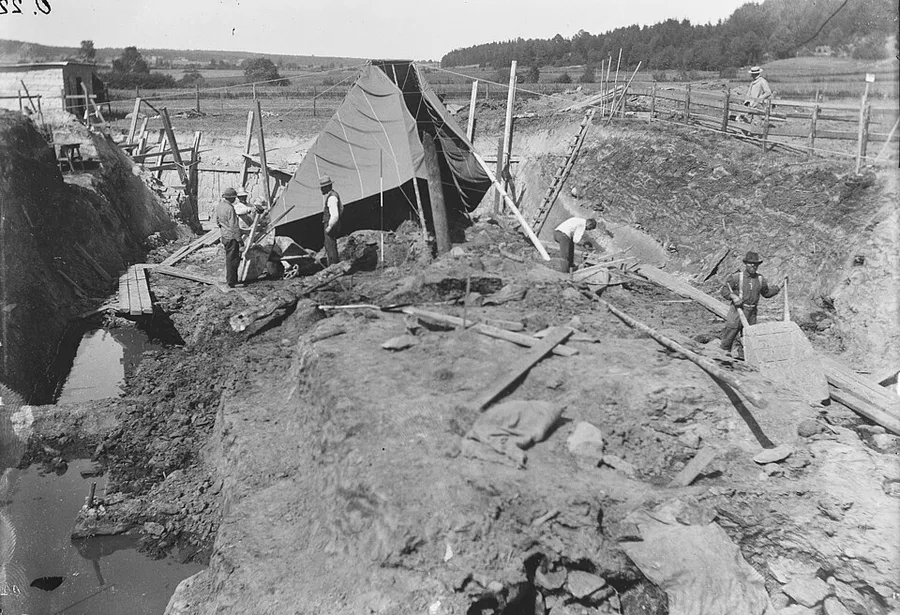 Black-and-white 1904 photograph of the Oseberg dig: the burial mound cut open into a deep pit, a dark canvas tent pitched over the find, workmen in hats standing on the raw earth, ladders and plank fencing around the excavation, farmland and forest beyond.