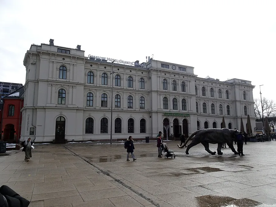 The historic 1882 East Station hall (Østbanehallen) on Oslo’s Jernbanetorget square — a pale neo-Renaissance building, now a hotel and the eastern face of Oslo Central Station — with the bronze tiger sculpture on the plaza.