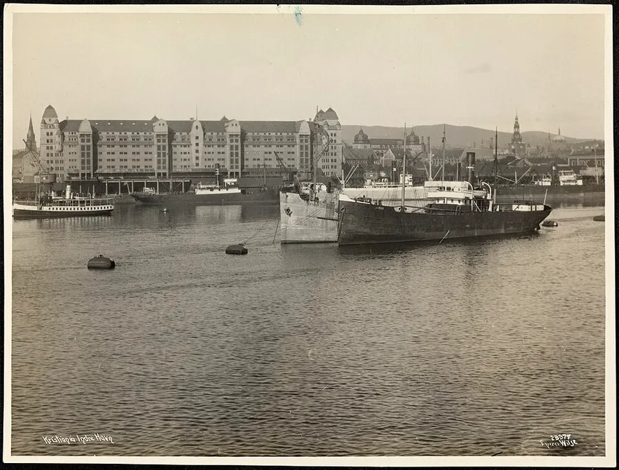 Black-and-white 1923 photograph of Kristiania's inner harbor at Bjørvika: a cargo steamer and small passenger boats moored on calm water, a long many-gabled harbor warehouse on the quay behind, and the city's spires and rooftops fading into a hazy hillside.