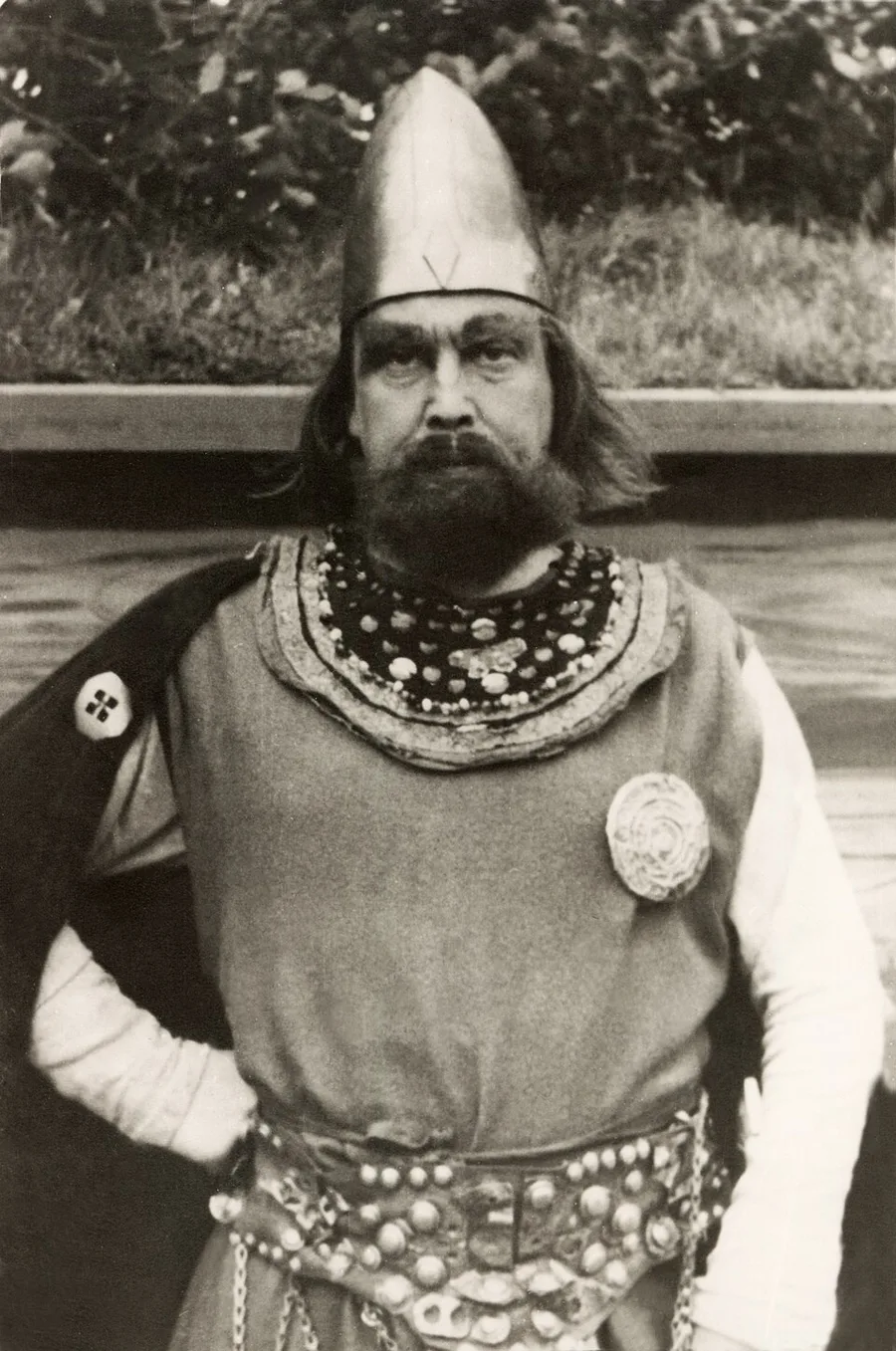 Black-and-white 1954 photograph from the first Spelet om Heilag Olav at Stiklestad: actor Gisle Straume costumed as King Olav — conical helmet, full beard, broad beaded collar, disc brooch and studded belt, dark cloak over one shoulder — before the open-air stage.