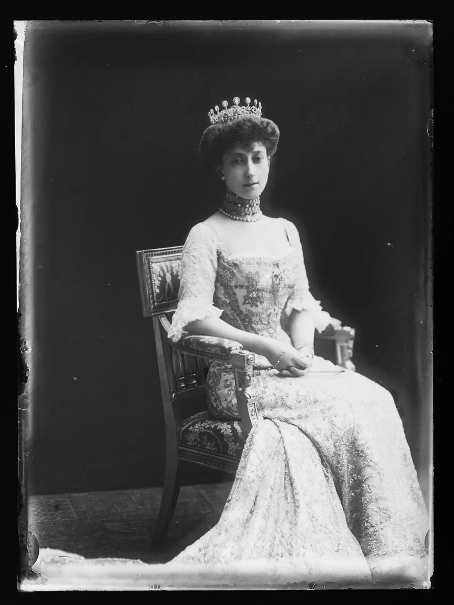 Glass-plate studio photograph of a young woman in an elaborate lace and jeweled gown with a pearl choker, seated in an ornate chair and wearing a diamond tiara — a 1906 portrait of Queen Maud of Norway.