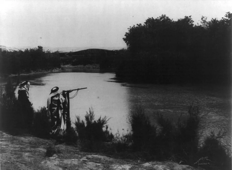 Early black-and-white photograph of the River Jordan curving between brush-covered banks, two robed figures standing on the near shore at left and low hills on the far horizon.
