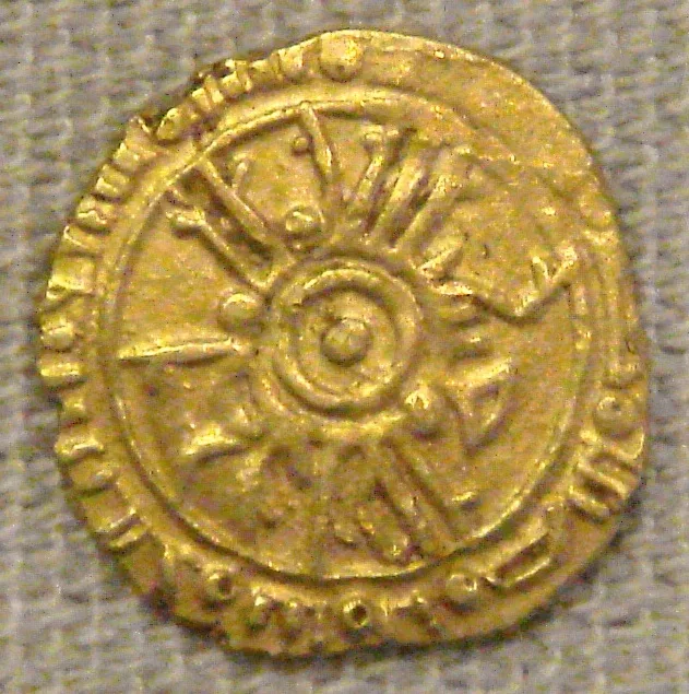 A small worn gold coin with a radiating wheel-like design around a central medallion, ringed by inscriptions in Arabic and Latin script, photographed against a gray fabric backdrop.