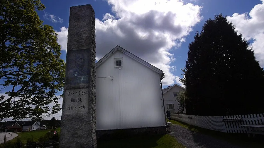 At the Hauge farm site on Rolvsøy: a rough granite memorial pillar bearing a portrait relief and the inscription 'Hans Nielsen Hauge 1771–1824' stands beside the white gable of the wooden museum building, with a picket fence, bench and trees around and houses in the distance.