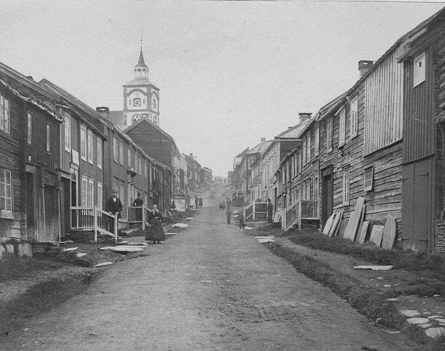 Black-and-white 1869 photograph looking up the unpaved main street of Røros, lined with dark timber-clad houses, a few pedestrians on the road, and the spired tower of the Bergstaden Ziir church rising at the street's end.