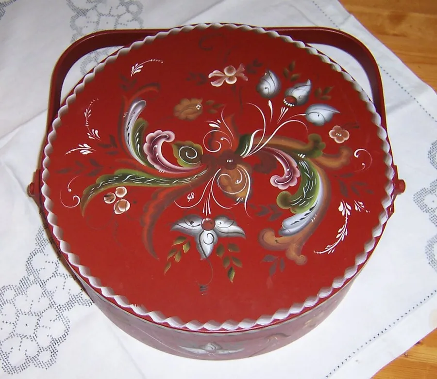 A round red lidded wooden tine with a scalloped rim, its lid hand-painted in rosemaling style with flowing floral scrollwork in green, pink, gold, and white, resting on a lace-edged cloth.