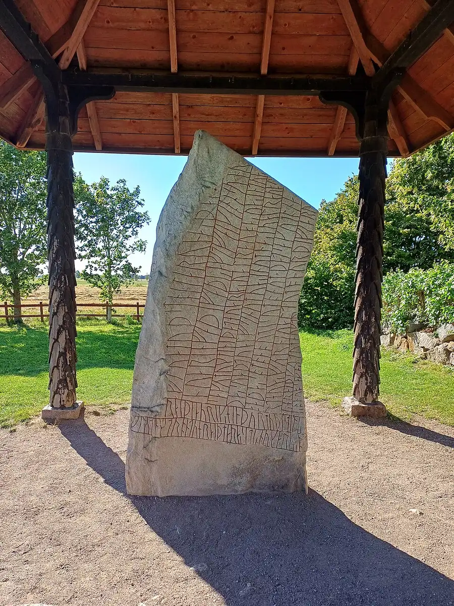 The Rök runestone, a tall granite slab densely covered edge to edge with carved runic inscription in vertical bands, standing outdoors under a peaked wooden shelter roof beside green fields and trees.