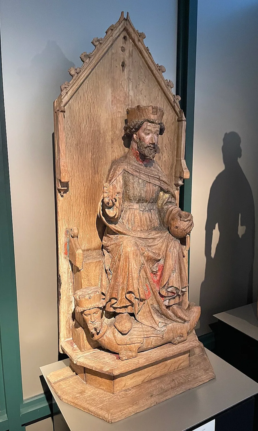 Medieval oak sculpture of Saint Olav enthroned within a pointed Gothic canopy: crowned and bearded, traces of red paint on his robes, a dragon-bodied creature with a crowned human head lying beneath his feet.