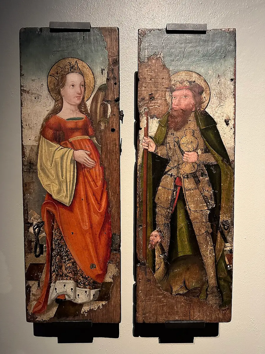 Painted wooden shrine doors: left, a crowned female saint in a red gown with a halo, hand resting at her waist; right, a bearded crowned king with a staff, standing over a small creature with a human head.