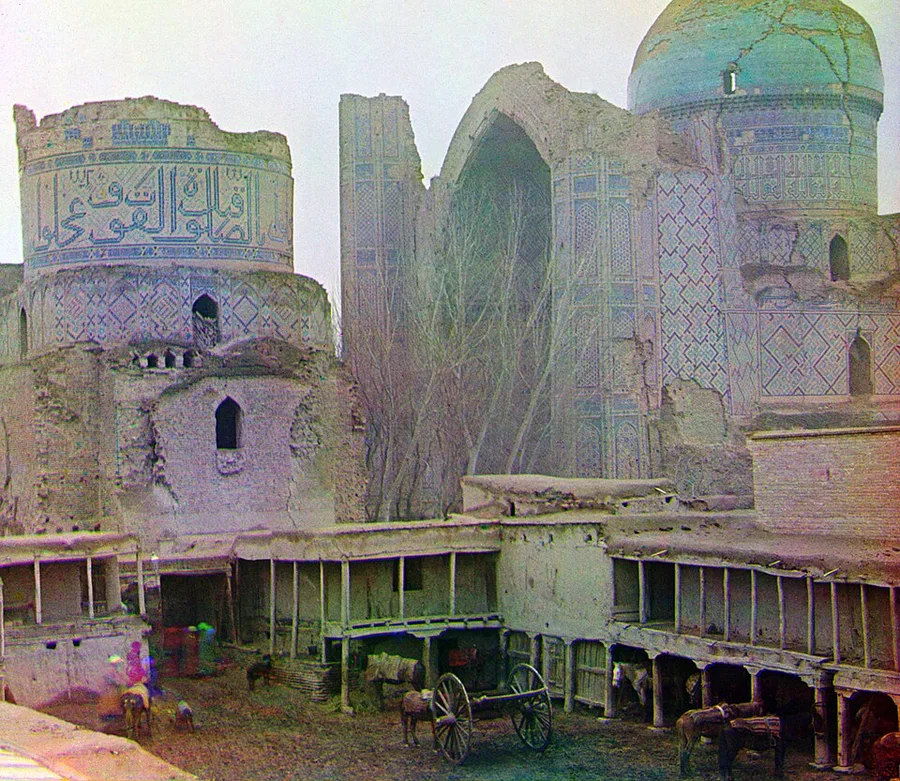 1907 hand-colored photograph of the ruined Bibi-Khanym Mosque in Samarkand: a crumbling turquoise dome and tiled portal rise behind a courtyard of arcaded ruins, bare trees, a horse-drawn cart, and tethered horses in the foreground.