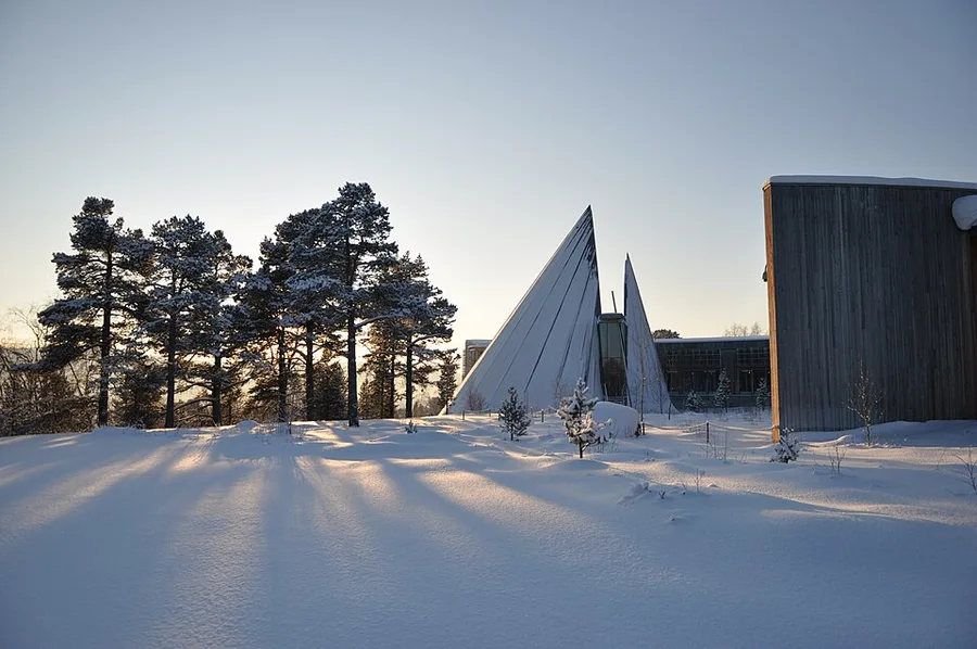 The Sametinget building in Karasjok at dusk: a tall, sail-like curved timber-clad plenary hall beside a dark wood-paneled administrative wing, snow-covered ground and pine trees in the foreground.