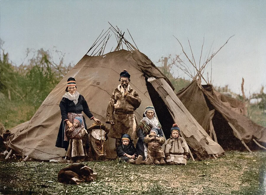 A hand-tinted photochrom of a Sámi family from around 1896 — adults and children in gákti dress gathered with their dog outside two lávvu tents.