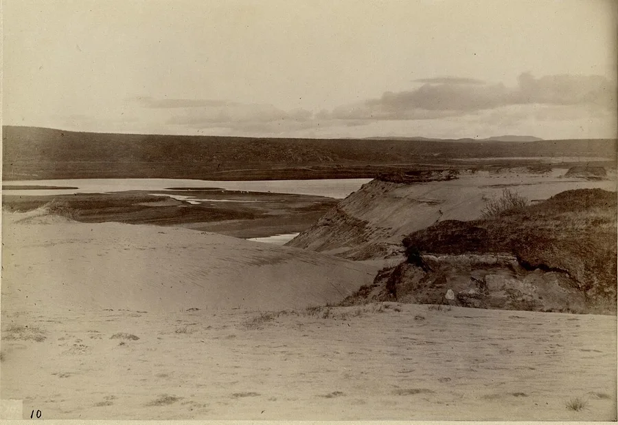 Sophus Tromholt's 1882–83 photograph of open country at Kautokeino — pale sand bluffs and dunes in the foreground, the braided river winding through the plain, and a low treeless fell horizon under a clouded sky.