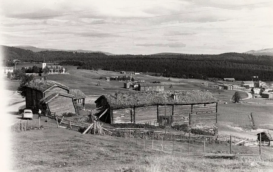 Black-and-white 1959 photograph: sod-roofed log farm buildings behind slanted wooden fences on a hillside at Oustad Søndre, with the Vingelen hamlet below — a white church tower among trees, scattered farms and hayfields — and forested ridges under a wide sky.