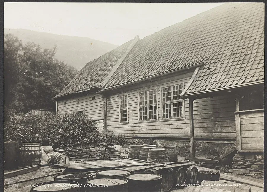 Black-and-white nineteenth-century photograph of a tall wooden gabled Schøtstue hall at Dramshusen in Bergen, casks and barrels stacked in the yard before its shingled roof and rows of windows.