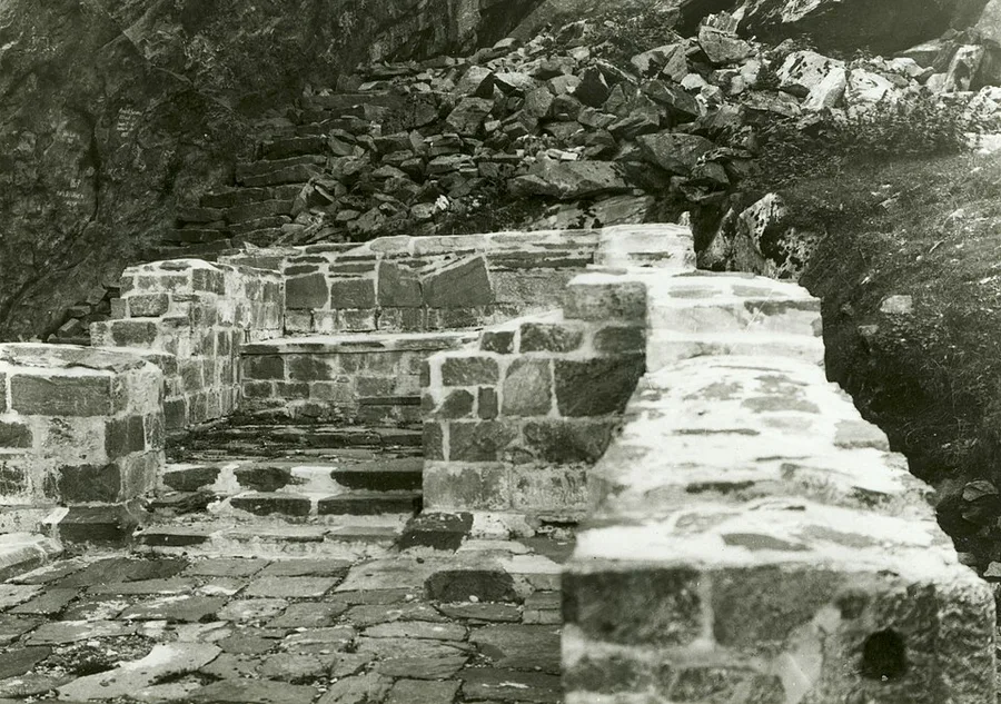 Black-and-white 1915 photograph of the low stone corner walls and steps of the Benedictine monastery ruins on Selja island, a rubble-covered slope rising behind.