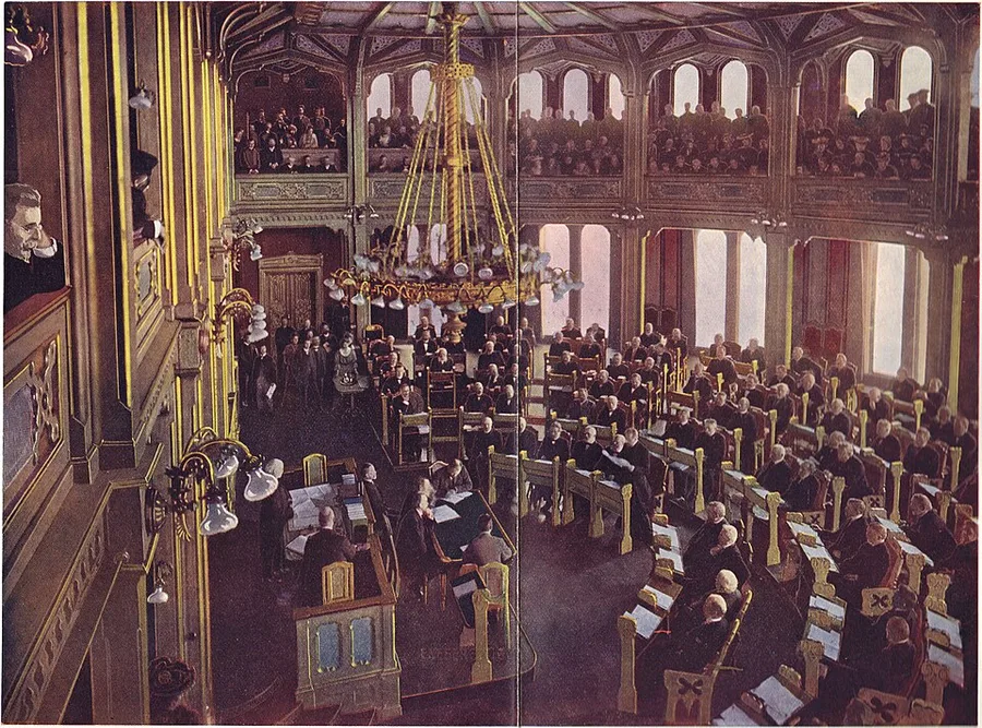 Hand-tinted photograph of the ornate Storting chamber packed with seated representatives and a crowded public gallery above, lit by a large brass chandelier — the 7 June 1905 session where the declaration dissolving the union was read.