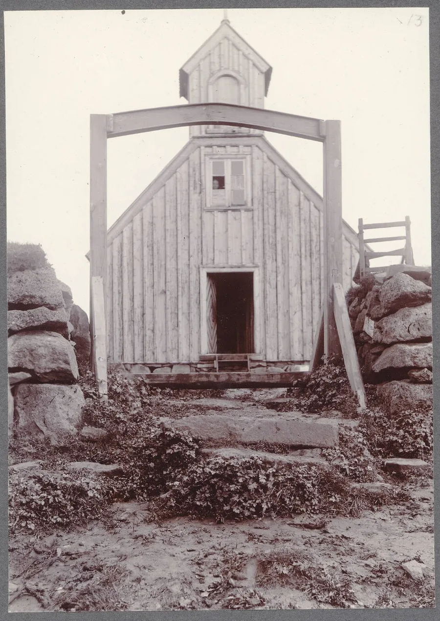 Circa-1900 photograph of the wooden church at Skálholt seen through its freestanding timber entrance-gate frame, with stone steps leading up to the open doorway beneath the bell tower.