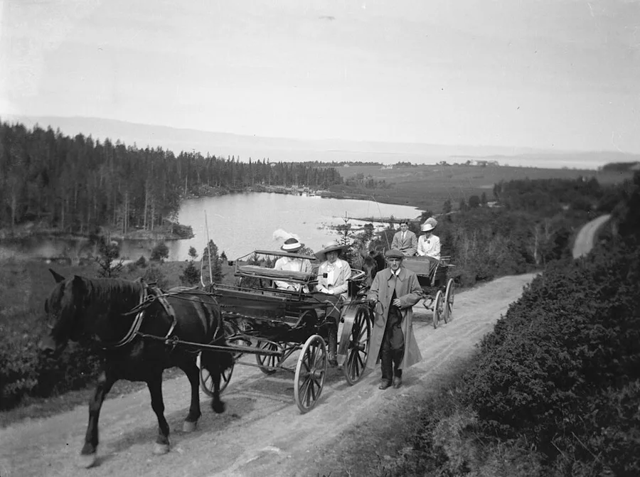A black-and-white ca. 1910 photo of two carriages paused on a country road above a lake, a single horse visible at the lead, well-dressed passengers in hats, and a coated man standing in the road beside the lead carriage.