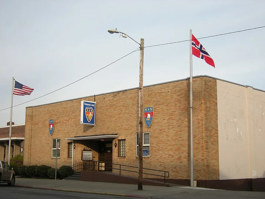 A single-story brick lodge building with a 'Normanna Lodge' banner and two Sons of Norway shield emblems on the facade, an American flag and a Norwegian flag flying from poles out front, photographed from the street.