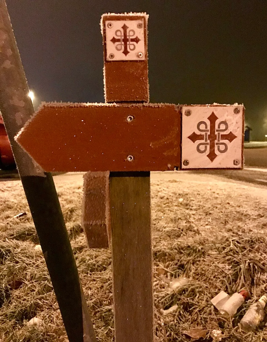 A frost-covered wooden waymark post at night: two square white plaques bearing the red St. Olav's cross, one atop the post and one beside a rust-red directional arrow pointing left, frosted grass and darkness behind.