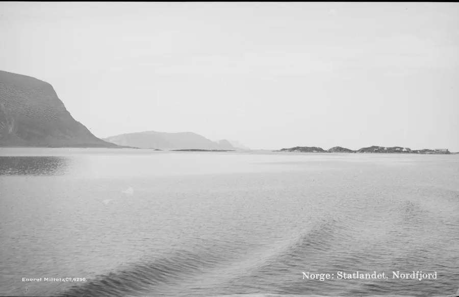 Photograph from about 1900, taken over a ship's wake off Stadlandet — the headland's dark cliff plateau dropping to the sea at left, low islets with a scatter of buildings to the right under a pale sky. 'Norge: Statlandet, Nordfjord' is printed in the negative.