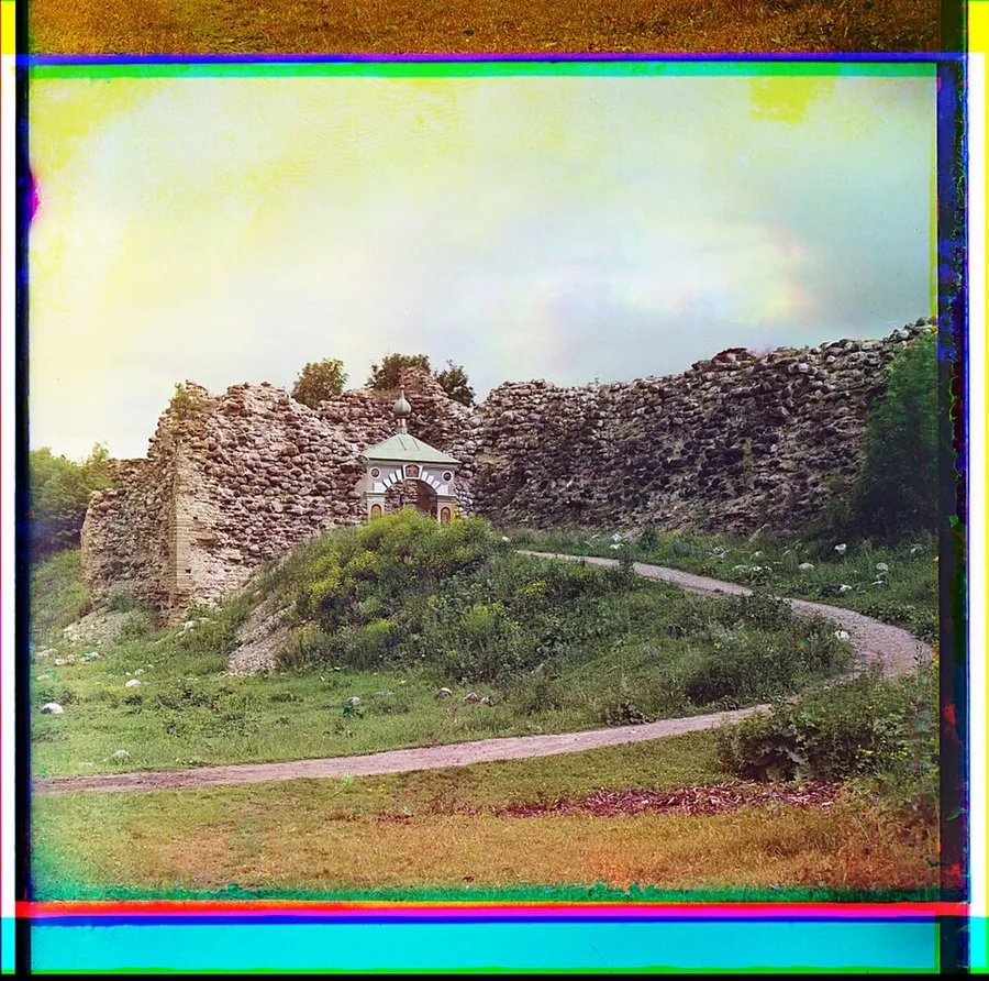 Early colour photograph, 1909: the ruined rubble walls of the Staraya Ladoga fortress rise over grassy mounds, a small white gate chapel with green roof and tiny dome set into them, dirt paths curving past, the plate edged with colour-fringed borders.