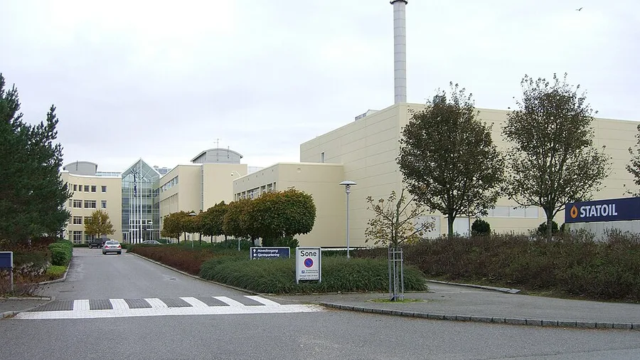 Daytime exterior photo of a low-rise cream-colored Statoil office building at Lura near Sandnes, with a tall industrial flue tower, glass entrance atrium, parking, and a blue Statoil sign in the foreground.