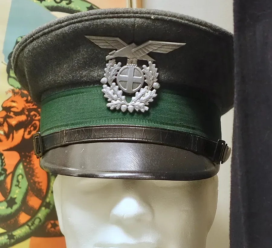 Close-up photograph of a grey Statspolitiet visor cap with a green band and a silver eagle-and-wreath badge over a cross emblem, displayed on a mannequin head.