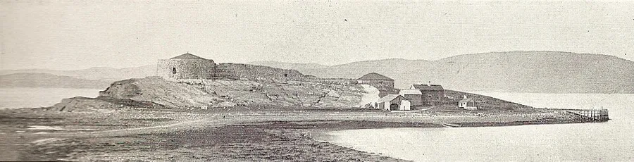 Wide, letterboxed sepia panorama of Steinvikholm Castle's ruined stone walls and round tower rising from a low tidal island, with a jetty and small outbuildings beside it and hills across the fjord in the distance.