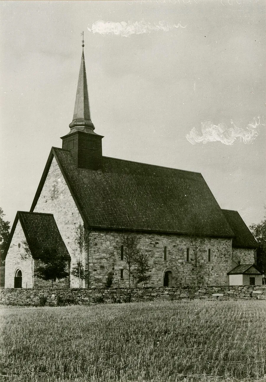 Black-and-white 1930 photograph of Stiklestad Church: a whitewashed medieval stone church with a tall dark spire and steep pitched roof, standing alone in open farmland behind a low stone wall.