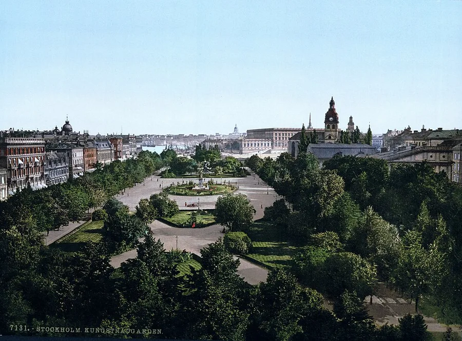 Color photochrom print of Kungsträdgården park in central Stockholm around 1900: tree-lined gravel walks, a central fountain and flower beds, with period buildings and a clock tower framing the far end of the square.