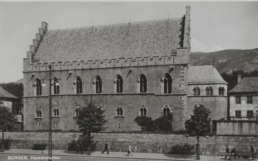 Black-and-white postcard photograph of Håkonshallen in Bergen: a long stone hall with stepped gables, crenellated parapet and tall Gothic windows rising above a walled street where a few pedestrians pass; printed caption 'Bergen. Haakonshallen.'