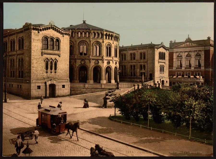 Tinted photochrome print of the Storting building in 1890s Kristiania, its ornate brick-and-stone facade seen across a plaza with a horse-drawn tram, pedestrians, and trees.