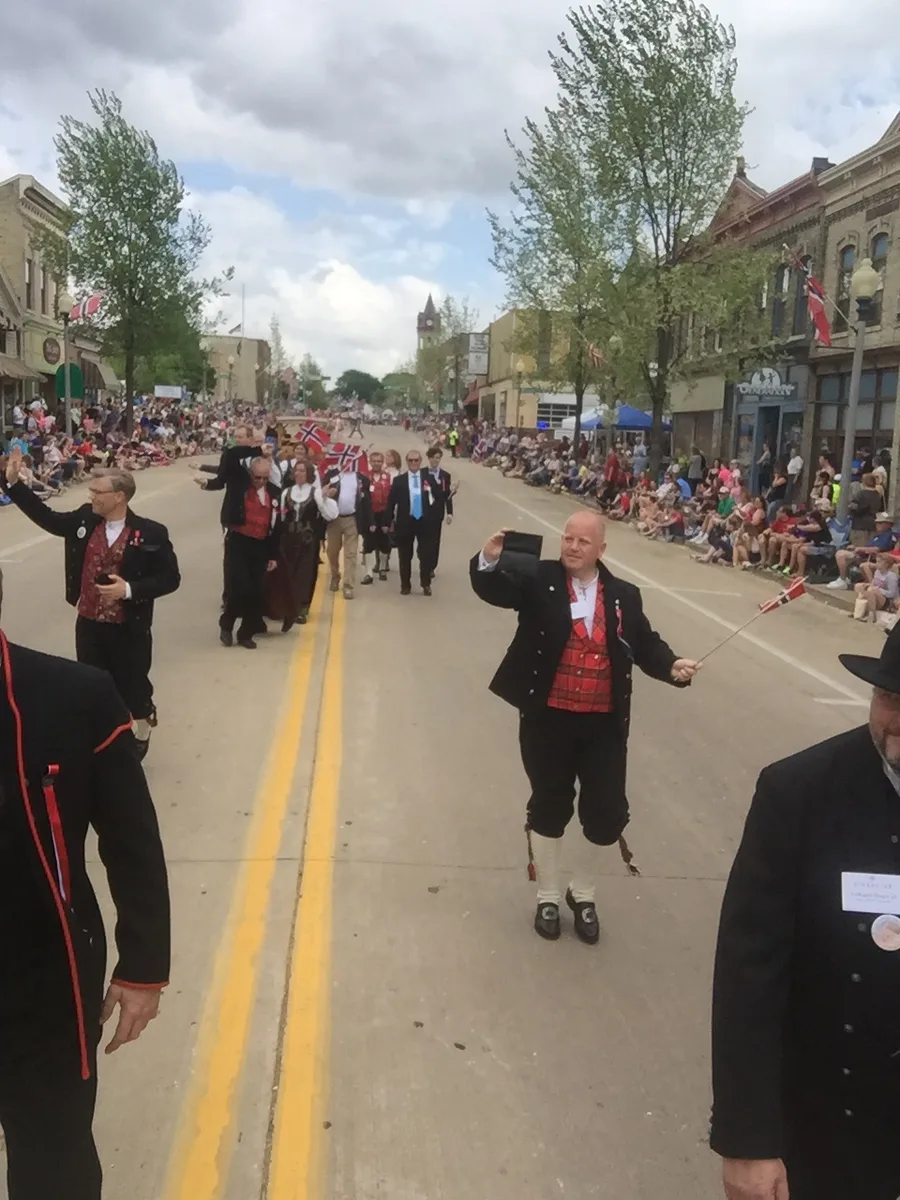 Marchers in black-and-red Norwegian bunad-style dress wave small Norwegian flags down Stoughton's main street during the 2015 Syttende mai parade, with spectators lining both curbs, brick storefronts hung with Norwegian flags, and a clock tower in the distance.
