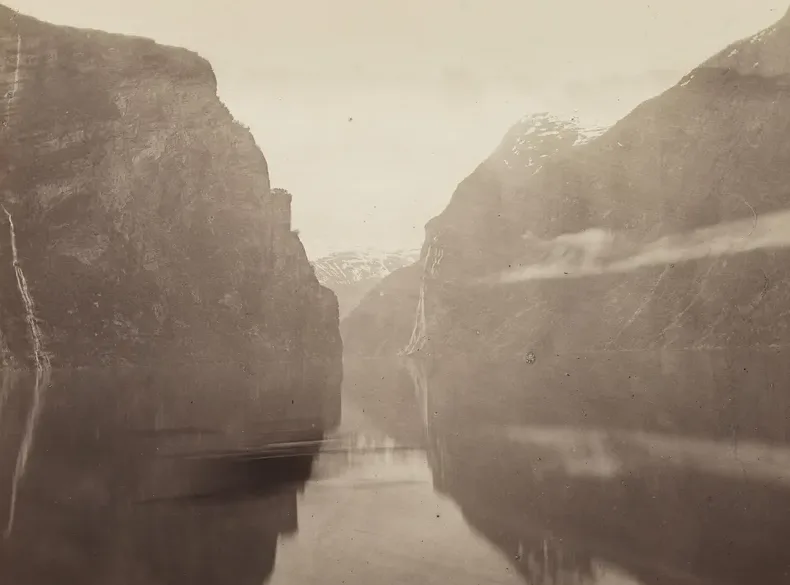 Knud Knudsen's nineteenth-century photograph of the Geirangerfjord in Sunnmøre — sheer fjord walls dropping to mirror-still water, thin waterfalls threading the cliffs, snow on the peaks at the fjord's head.