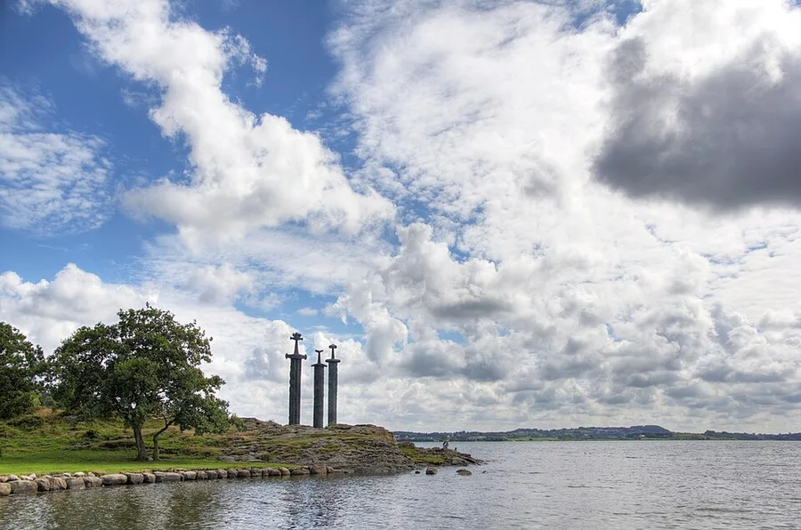 Fritz Røed's 1983 monument Sverd i fjell — three giant bronze swords driven point-down into the rock on the shore of Hafrsfjord, under a towering summer sky.