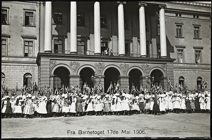 Black-and-white postcard photo captioned 'Fra Barnetoget 17de Mai 1906': rows of children in early-1900s dress and hats carry small Norwegian flags past the colonnaded facade of the Royal Palace in Christiania.